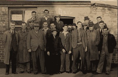 Sepia-toned photograph of 20 men standing in front of a brick building