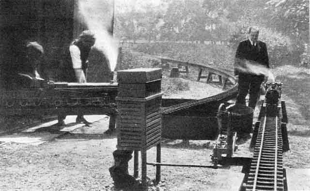 A man observing a model steam locomotive on a raised outdoor track, while two men work on another locomotive