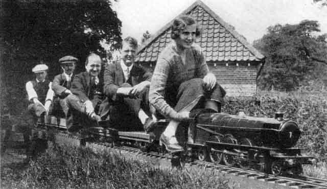 A woman and four men riding a model steam train past a brick building