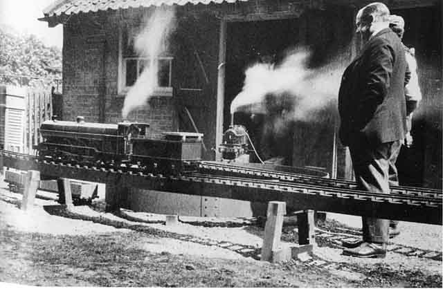Two men observing a model steam locomotive on a raised outdoor track, while another man stands next to a second locomotive in the doorway of a brick building