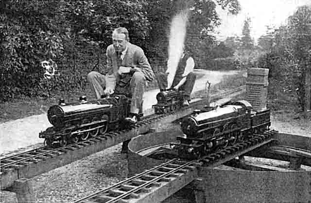 Two men riding model steam locomotives past another locomotive on an outdoor turntable