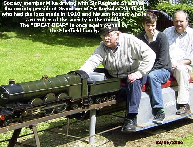 Three men riding a model steam locomotive on a raised outdoor track, dated ‘02/06/2006’ and captioned ‘Society member Mike driving with Sir Reginald Sheffield, the society president Grandson of Sir Berkeley Sheffield who had the loco made in 1910 and his son Robert who is a member of the society in the middle. The “GREAT BEAR” is once again owned by The Sheffield family.’